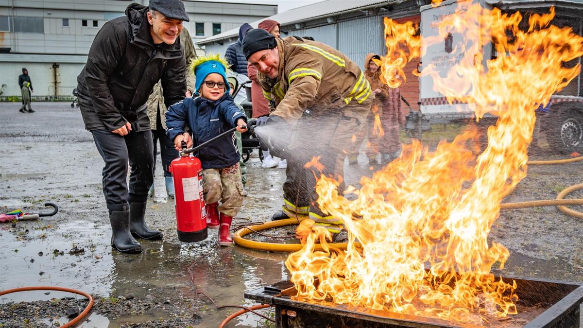 Far og barn med slokkeapparat sammen med brannkonstabel og flammer - Klikk for stort bilde